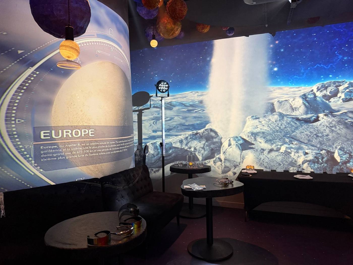 Coin salon devant une projection de la lune Europe, avec lunettes futuristes sur les tables.