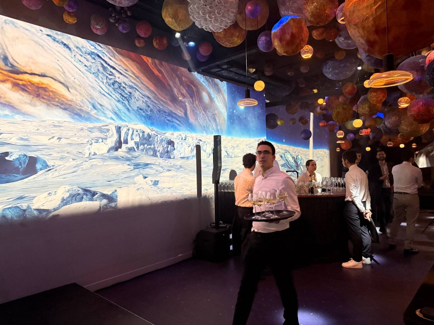 Salle sur le thème de l'espace : projection de paysage glacé sur le mur, planètes suspendues au plafond, serveur avec plateau de verres et bar rond.