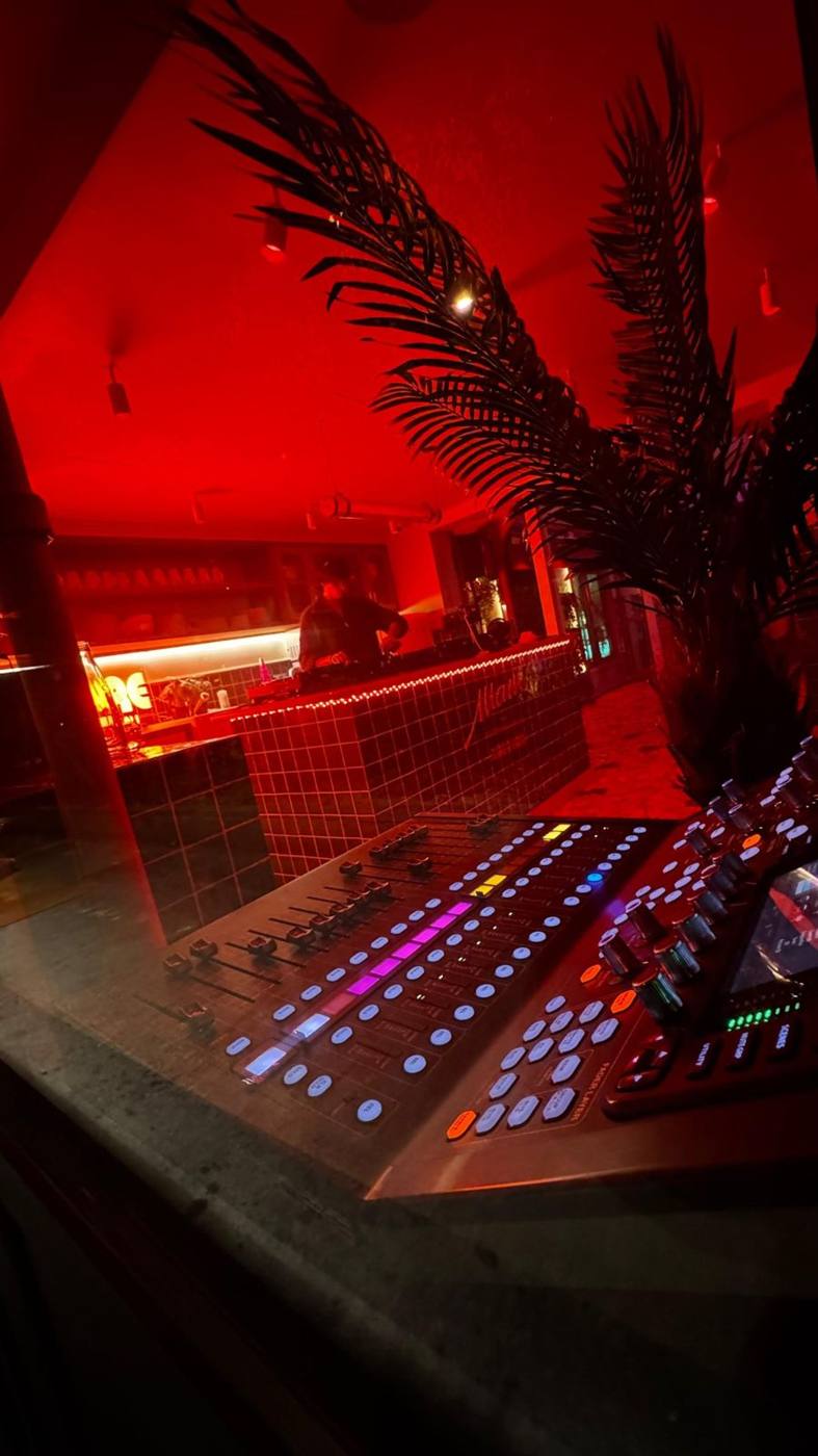 Console de mixage numérique au premier plan, DJ et palmier en arrière-plan sous éclairage rouge.