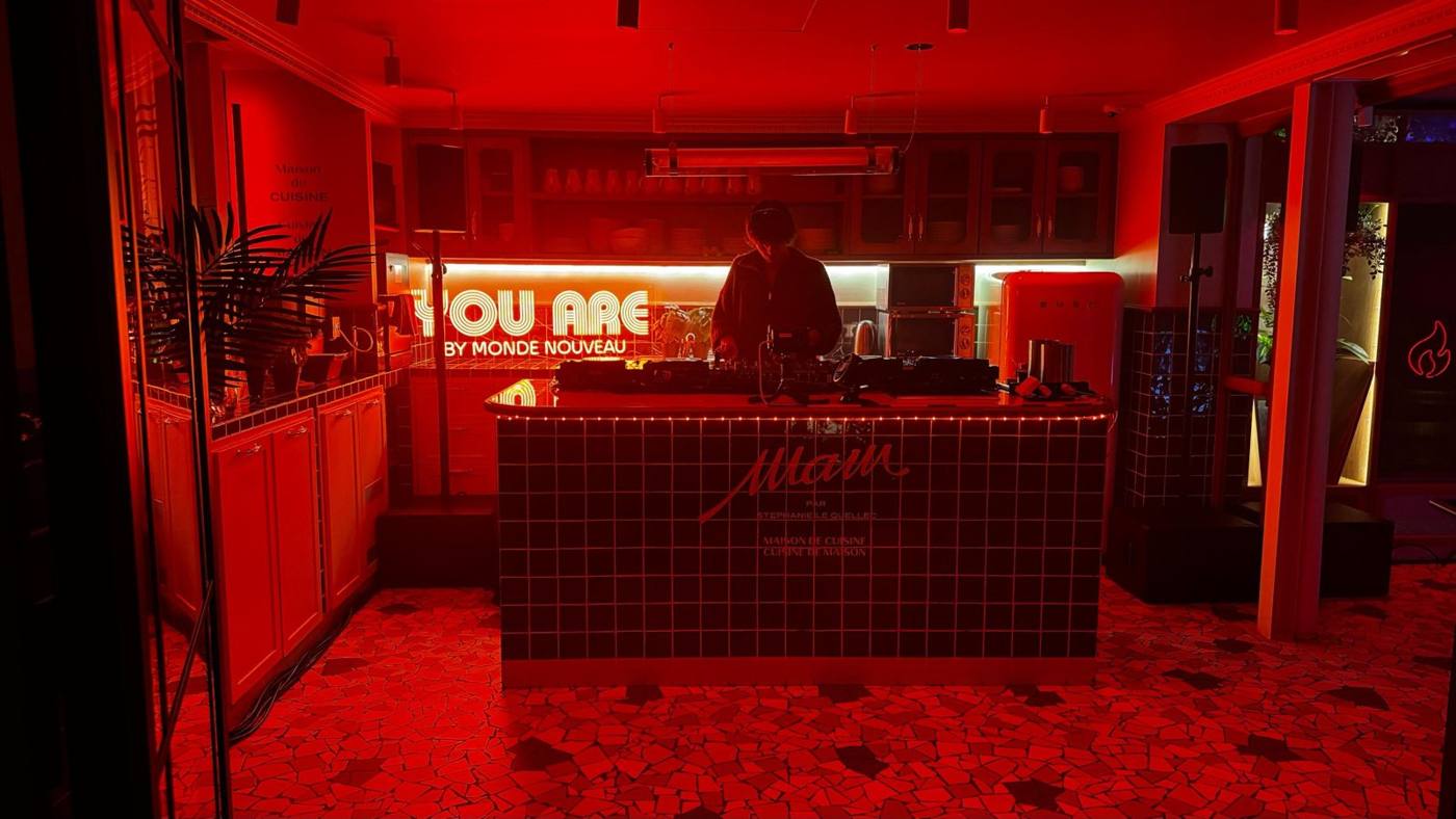 DJ derrière un îlot carrelé dans une cuisine éclairée en rouge, néon « You are by Monde Nouveau » au fond, enceintes sur pieds.