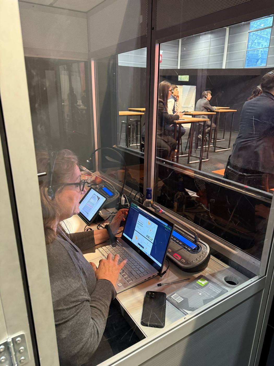 Interprète au travail dans une cabine vitrée, ordinateur et pupitre d'interprétation.