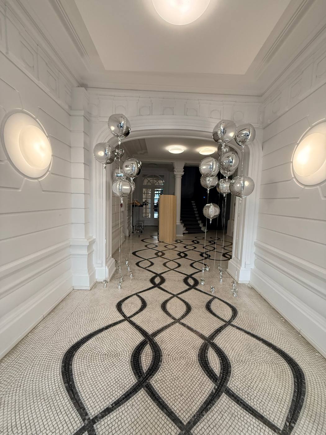 Hall d'immeuble au sol en mosaïque noir et blanc, ballons argentés en arche.