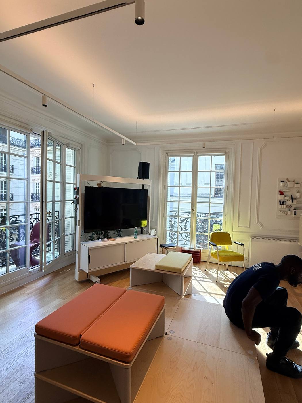 Salon de l'appartement-showroom : écran sur meuble mobile, assises orange et jaune, technicien accroupi, portes-fenêtres sur balcon.